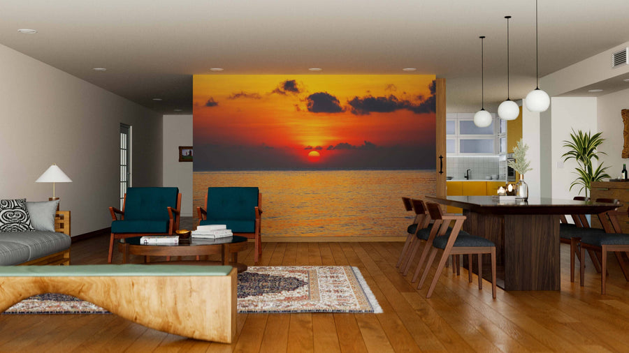 Modern living and dining room with wooden floors and furniture, featuring the Sea Sunset Deep Red Wall Mural depicting a vivid orange sunset over the ocean. The space has teal armchairs, a bench, rugs, and globe pendant lights above the dining table.