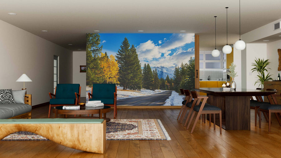 A modern living and dining room features wood floors, stylish furniture, and a Snowy Autumn Colorado Mountains Road Wall Mural. Warm lights, rugs, and pendant lamps add coziness, with the kitchen visible in the background.