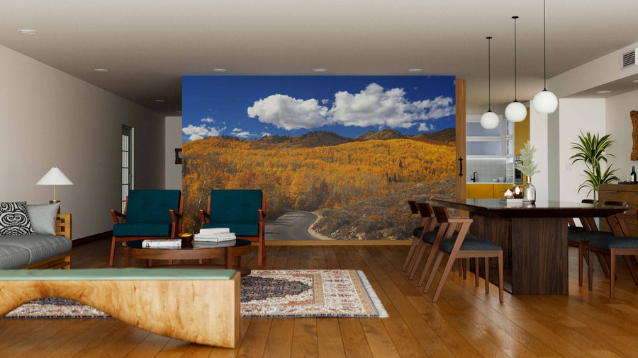 A modern living and dining area with wood floors, teal armchairs, and round pendant lights features the Wasatch Mountain State Park Wall Mural, showcasing golden autumn trees beneath a clear blue sky.