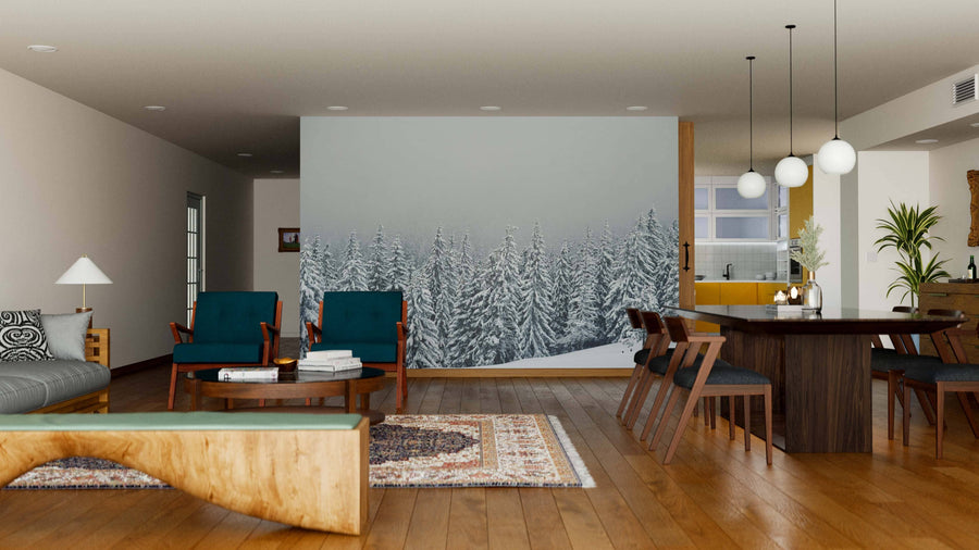 A modern living-dining area features wood floors, teal armchairs, a gray sofa, a wooden dining table, and pendant lighting. The back wall displays the "Winter Trees on Mountain Wall Mural," adding a cozy touch to the minimal space.