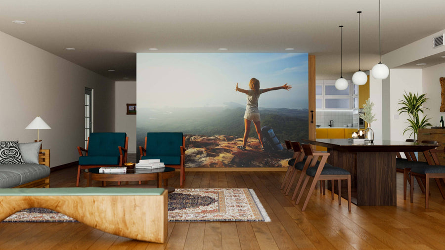 A modern living-dining area with wood floors, teal chairs, and pendant lights features the Woman Backpacker Wall Mural—a peel and stick mural of a woman on a mountaintop, arms outstretched, gazing over a scenic valley at sunset.