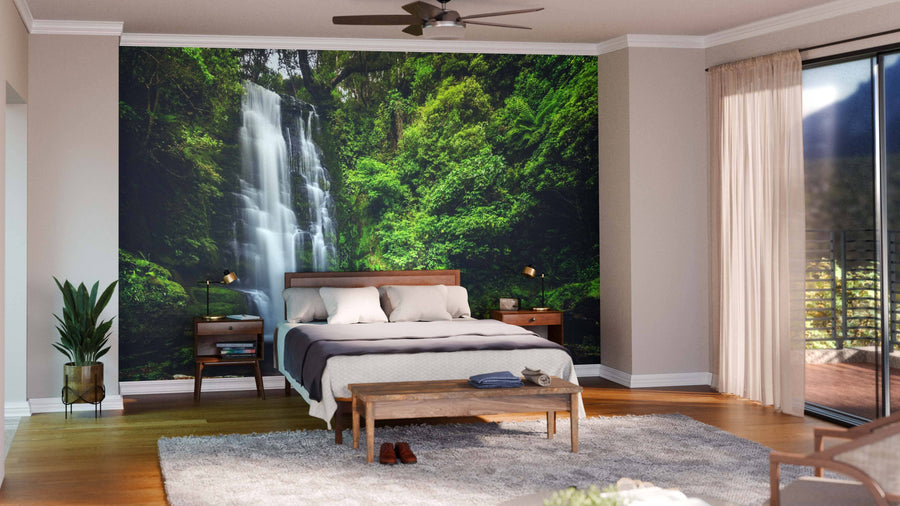 A modern living and dining area with wood floors, mid-century furniture, and a Maclean Falls Wall Mural depicting a lush green forest and waterfall. The space includes blue chairs, a wooden table, floor rug, and hanging globe lights.