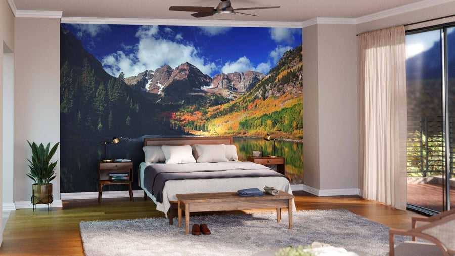 A modern living-dining room with wooden floors and contemporary furniture features the Maroon Bell Mountains Wall Mural, a large peel-and-stick design depicting a scenic mountain lake in autumn, adding depth and rich color to the warm, neutral decor.
