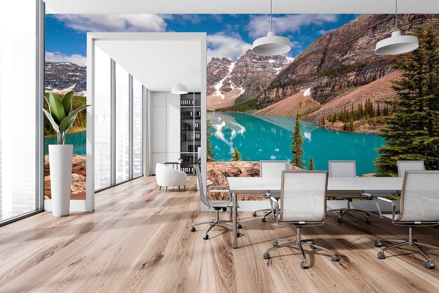 A modern bedroom features a large bed, potted plants, and a wooden bench. Sunlight warms the space through glass doors, highlighting the Mt. Allen and Moraine Lake Wall Mural of turquoise water and mountains behind the bed.