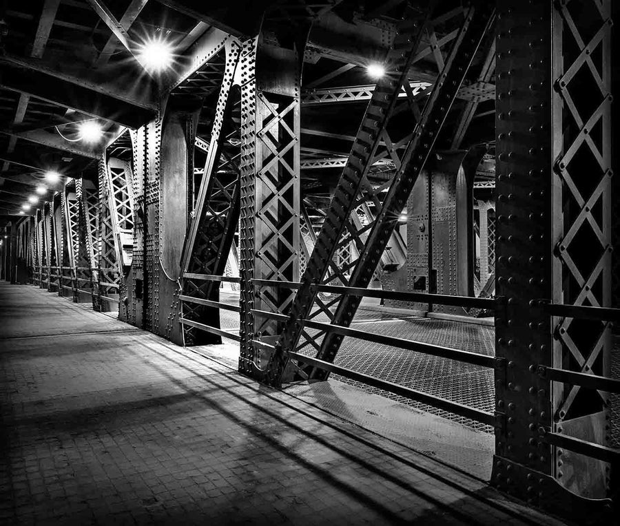 The Chicago Bridge Wall Mural depicts a dimly lit black and white scene of a metal bridge interior with crisscrossing beams, riveted columns, and geometric patterns that cast dramatic shadows on the concrete walkway, all softly illuminated by overhead lights.