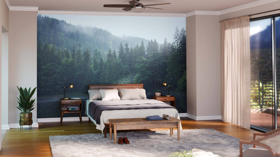 A modern bedroom features a large Morning on the Mountain Lake Wall Mural, a wooden bed with white and gray bedding, two nightstands with lamps, a plant, a bench, a gray rug, and sliding glass doors with beige curtains letting in natural light.