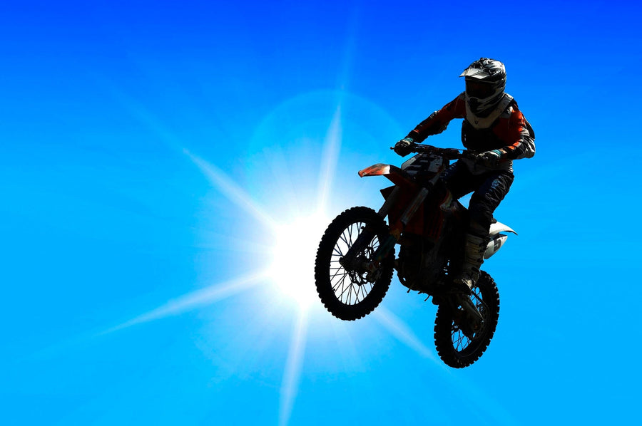 A clean garage with black cabinets and neat tools features the Motocross Wall Mural showing a rider against a bright blue sky. A classic motorcycle is parked inside, and a garden hose hangs by the window overlooking outdoor plants.