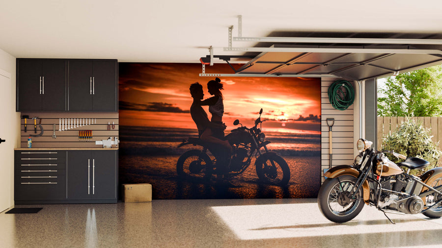 A tidy garage showcases dark cabinets and a vintage motorcycle. The back wall features the Beach Riders Wall Mural, showing two people silhouetted on a motorcycle at sunset by the beach. Tools are neatly arranged, sunlight comes in from the right.