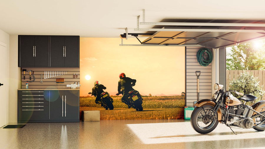 A tidy garage features black cabinets and neatly arranged tools on the left, a vintage motorcycle on the right, and an open wall adorned with the Biking on Asphalt Wall Mural, showcasing two riders at sunset across a grassy field as sunlight brightens the space.