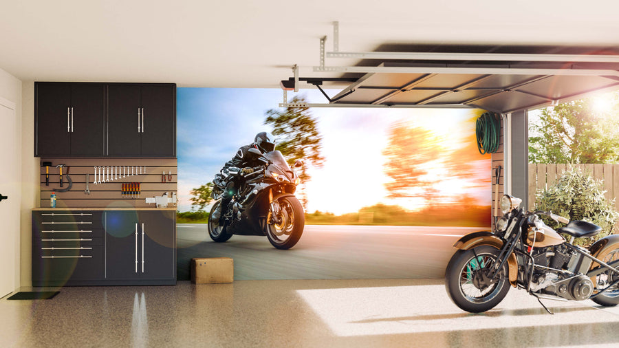 A modern garage with sleek black cabinets and organized tools features the Blazing Through Roads Wall Mural, showing a sport motorcycle speeding by. Sunlight through the open door highlights a classic bike inside and greenery outside.