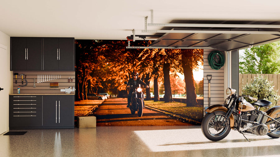 A modern garage with dark cabinets, organized tools, and a classic motorcycle inside. The back wall features the Cafe Racer Motorcycle Wall Mural, showing a rider cruising down a tree-lined street with autumn leaves for a warm, seasonal vibe.