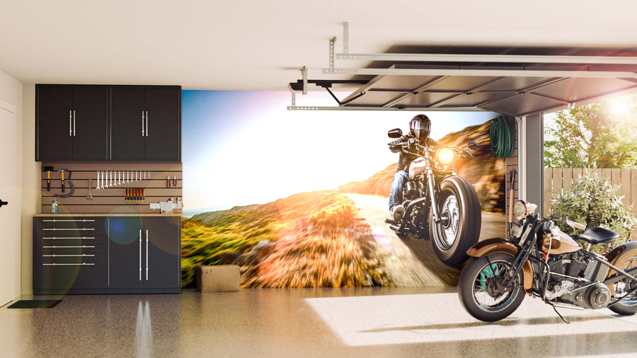 A modern garage features sleek black cabinets and organized tools on the left. A motorcycle sits on the glossy floor, while a Coastal Highway Ride Wall Mural of a rider on a scenic road decorates the wall behind. The garage door is partially open.