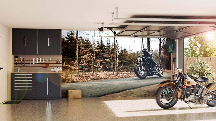 A clean, modern garage with black cabinets and neat tools on the left. A classic motorcycle rests on a polished floor. The back wall displays the Forest Motorcycle Ride Wall Mural showing a motorcyclist speeding down a sunlit forest road.