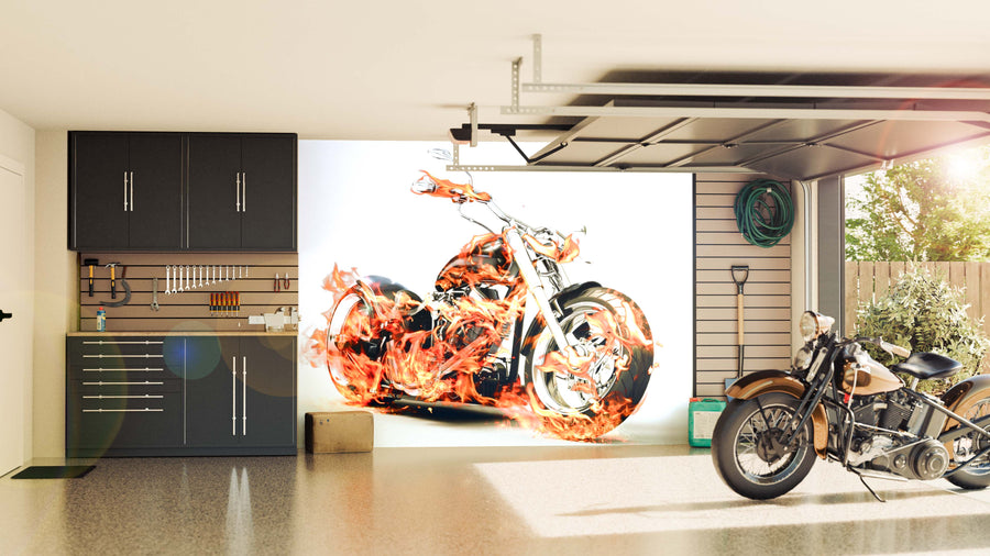 A modern garage with dark cabinets and organized tools features the Burning Bike with Flames Wall Mural, showing a vivid flaming motorcycle. Sunlight streams in through the open door, revealing greenery outside and garden equipment neatly stored.
