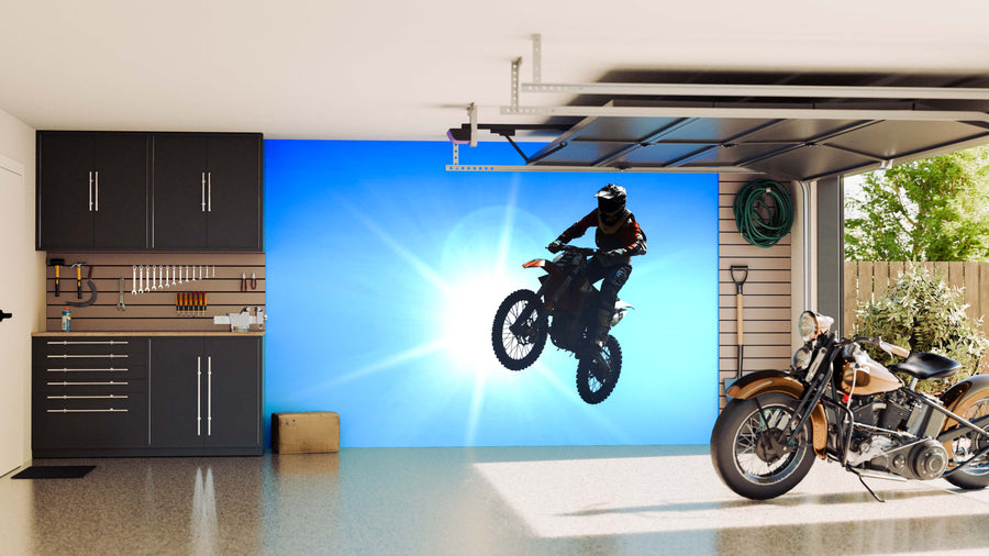 A clean garage with black cabinets and neat tools features the Motocross Wall Mural showing a rider against a bright blue sky. A classic motorcycle is parked inside, and a garden hose hangs by the window overlooking outdoor plants.