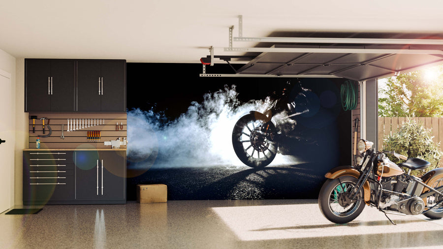 A modern garage with black cabinets, neatly arranged tools, and a classic motorcycle features the Motorcycle Burnout Smoke Wall Mural on the back wall, showing smoke and sunlight pouring in through the open garage door.