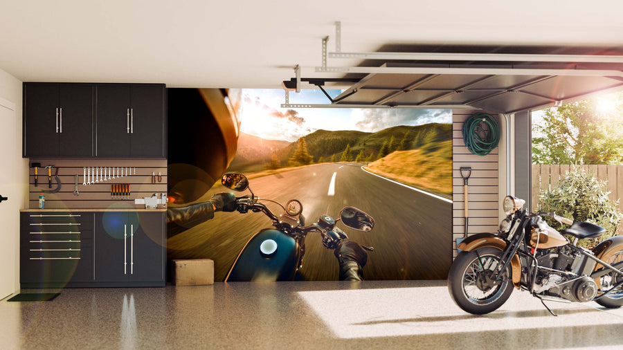 A sleek, organized garage with dark cabinets, tools, and a motorcycle. The back wall showcases the Mountain Drive Wall Mural—a peel and stick mural of a rider’s view on an open mountain road—illuminated by natural light from the open door.