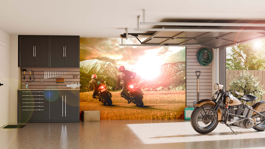 A tidy garage with dark cabinets, organized tools on the wall, and a vintage motorcycle features sunlight streaming through a window and the vibrant Mountain Ride Wall Mural, depicting two motorcyclists riding into a sunlit landscape.