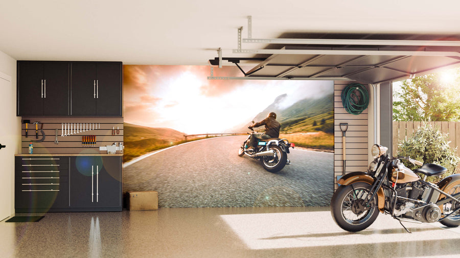 A tidy garage with black cabinets, tools neatly hung, and a brown motorcycle inside. One wall features the Mountain Road Escape Wall Mural showing a person riding a motorcycle on a curving road at sunset, with coiled hoses hanging nearby.
