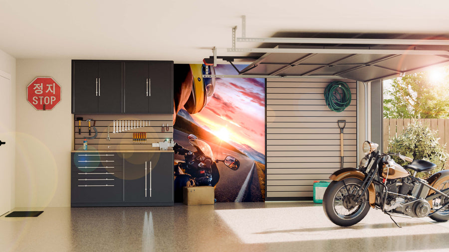A modern garage with gray cabinets, a wall shelf holding tools, a coiled green hose, and a vintage motorcycle. The back wall showcases the Rider at Sunset Wall Mural, glowing in sunlight from the open garage door.