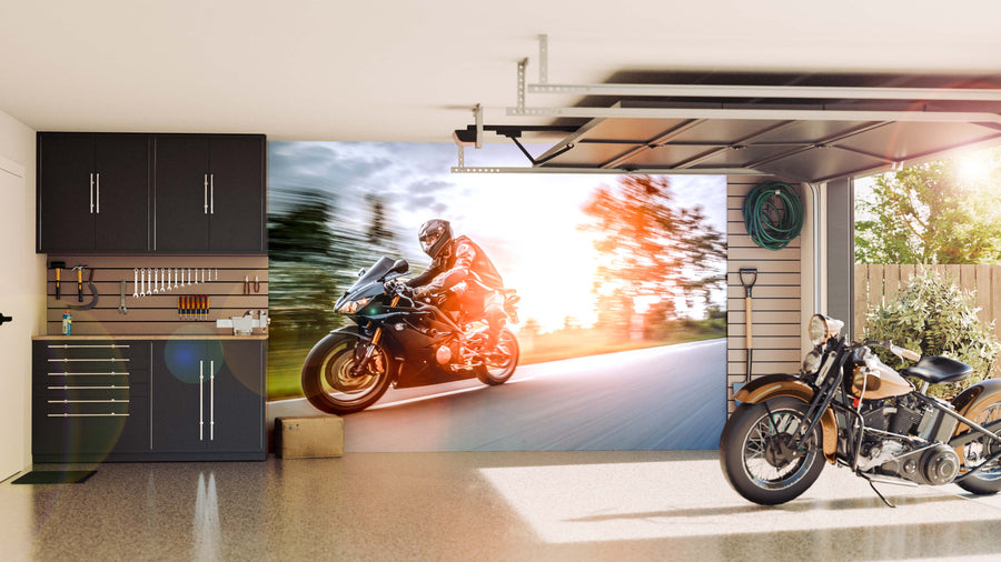 A modern garage features sleek cabinets and tools on the left, a “Riding into the Sunset Wall Mural” of a motorcycle racer on the back wall, a vintage motorcycle on an epoxy floor, and sunlight streaming through an open door to the right.