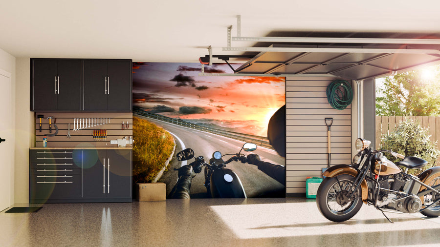 A garage with black cabinets, neatly organized tools, a shovel, a hose, and a retro motorcycle. The back wall showcases the Road Horizon Wall Mural depicting a person riding at sunset as sunlight streams through the open door.