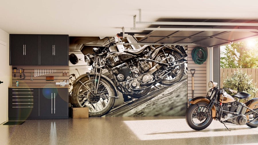 A modern garage with sleek black cabinets and tool storage showcases the Rustic Charm Wall Mural—a striking black-and-white vintage motorcycle design. Another vintage bike sits on the floor, sunlight pours in, and greenery is visible outside.