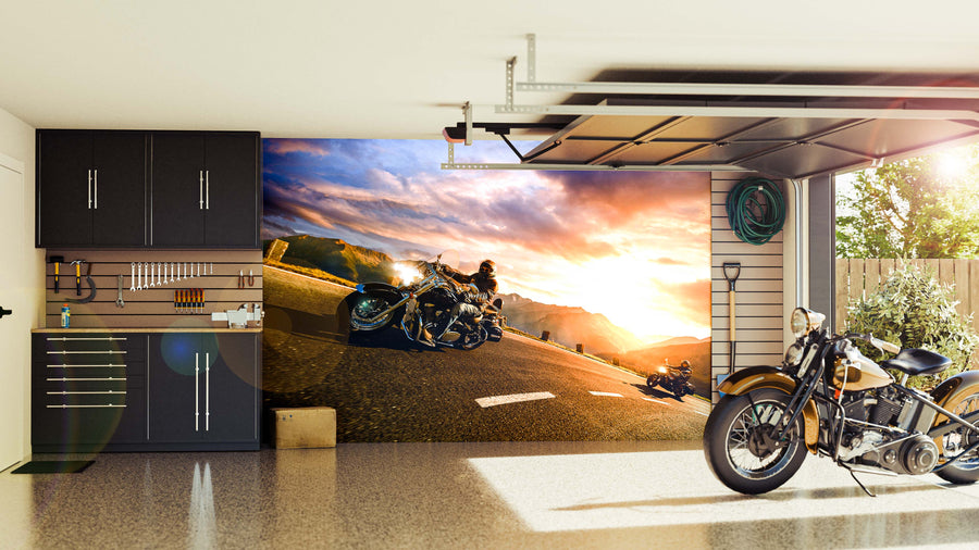 A modern garage with black cabinets, wall-mounted tools, and a retro motorcycle features the Sunset Ride Wall Mural on its back wall—depicting motorcyclists riding on a scenic mountain road at sunset.