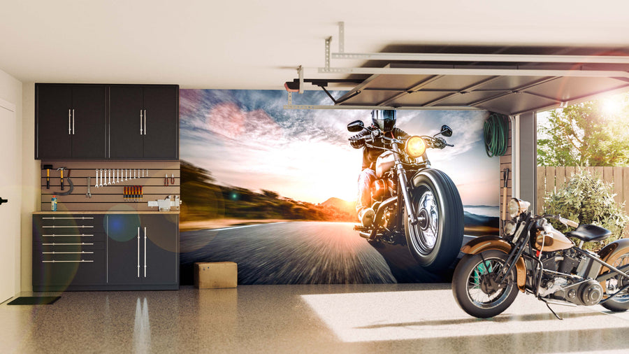 A tidy garage with black cabinets and organized tools, featuring the Sunset Ride Rush Wall Mural—a large peel and stick mural showing a motorcycle speeding down a road at sunset. A vintage-style motorcycle sits on the speckled floor near the open door.