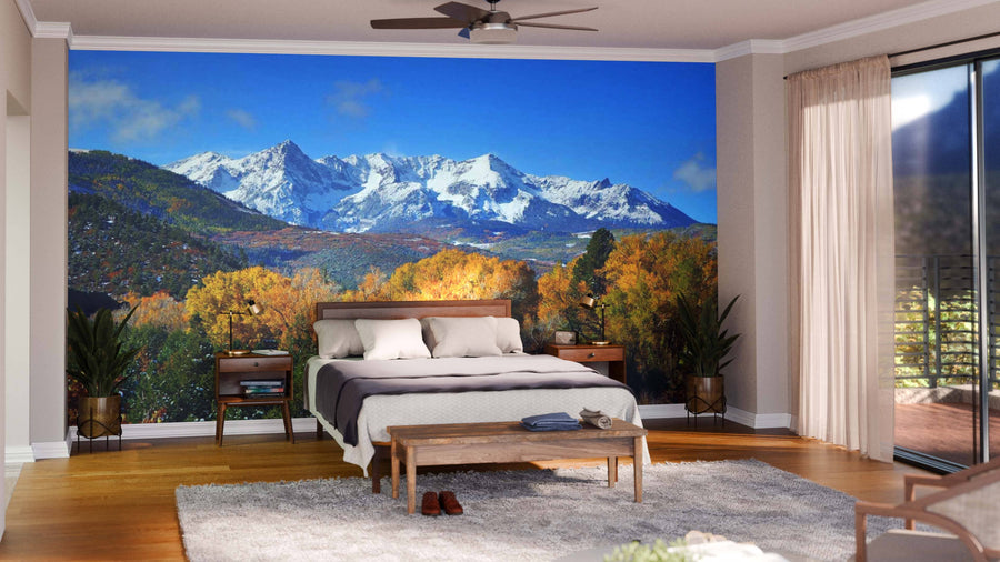 A modern living-dining room features wooden floors, green armchairs, a large dining table, and globe pendant lights. The Mount Sneffels Panoramic View Wall Mural displays snow-capped peaks above autumn trees as a stunning nature-inspired focal point.