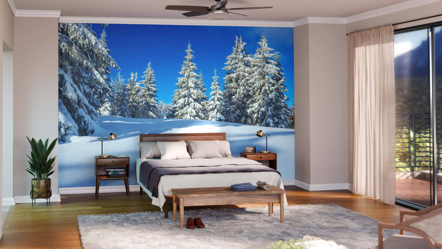 A bright, modern bedroom features a brown wood bed, two matching nightstands, plants, a rug, and a bench. Behind the bed is the Mountain Forest With Snow Wall Mural, showing snowy pine trees beneath a blue sky. Glass doors open to a sunny balcony.