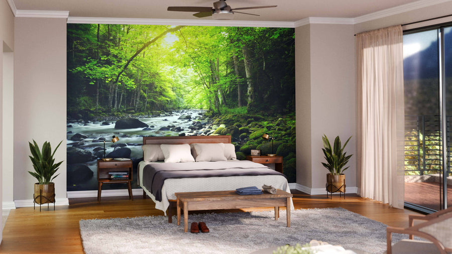 A modern living-dining space with wooden floors, blue and wood furniture, pendant lighting, and a large Mountain River in the Wood Wall Mural depicting a sunlit forest and stream for a serene, inviting ambiance.