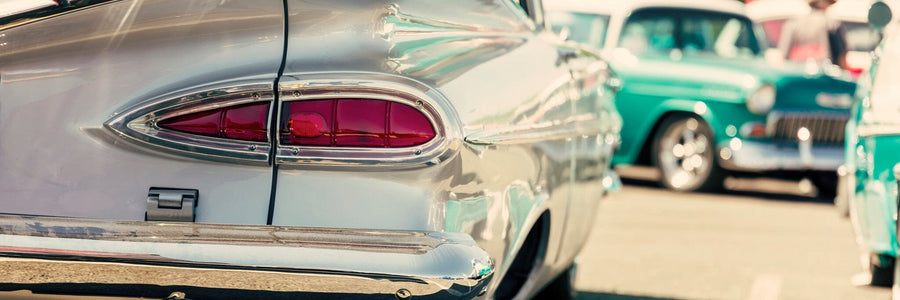 A modern bedroom features a desk, chair, and bed with yellow pillows. Behind the bed, the Old American Car Wall Mural with a shiny chrome tail fin and red tail light creates a vintage vibe. Sunlight streams through the blinds.