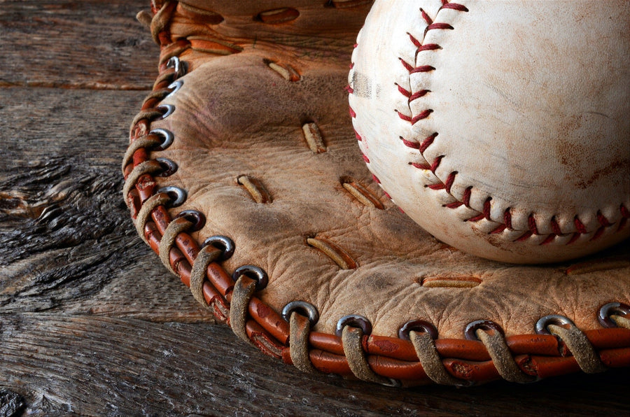 A modern bedroom centers around the Used Baseball Equipment Wall Mural, showing a baseball in an old glove. The space also has a desk with computer, bed, white cabinets, wall art, a soccer ball on the floor, and sunlight streaming through blinds.