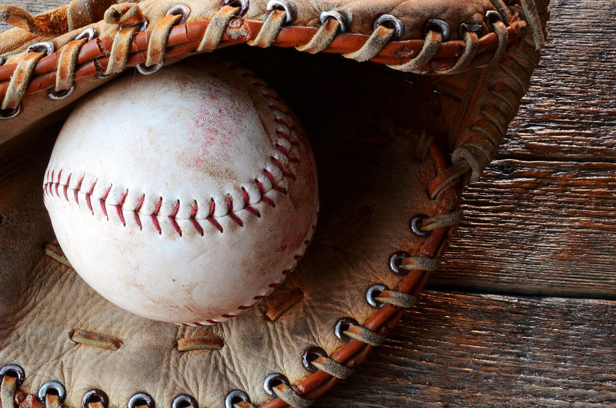 A modern bedroom with a bed, desk, computer, and chair features the Old Baseball Equipment Wall Mural—a close-up of a baseball in a worn glove on rustic wood. Sunlight shines through blinds, and a soccer ball rests near the bed.