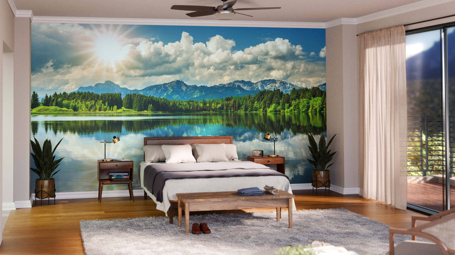 A modern bedroom with a bed, plants, and wooden furniture features the Panoramic Landscape with Lake Wall Mural depicting a sunny lake, trees, and mountains. Sunlight pours in through sheer curtains on glass doors, creating a serene atmosphere.