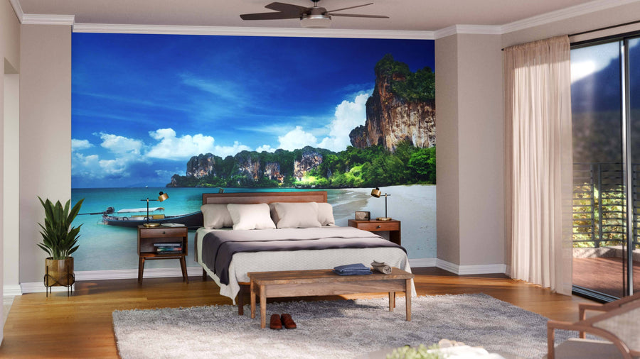 A modern living-dining room with wood floors and teal chairs features the “Railay Beach in Krabi Thailand Wall Mural,” depicting a tropical beach. Globe lights, a sofa, rugs, and natural decor complete this inviting space.