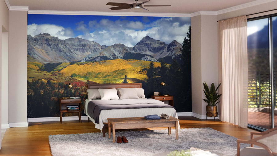 A modern bedroom features the San Juan Mountains Over Forest Wall Mural—a large, custom-printed peel and stick mural with grey mountains and green-yellow trees—behind the bed. Wood floors, mid-century furniture, plants, and sliding glass doors add to the serene vibe.