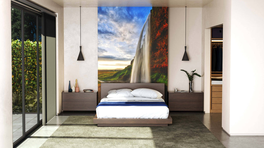 A modern living-dining room with mid-century furniture and wooden floors features the Seljalandfoss Waterfall Wall Mural on an accent wall, bringing natural beauty and a sunset landscape into the open, neutral-toned space with contemporary lighting.