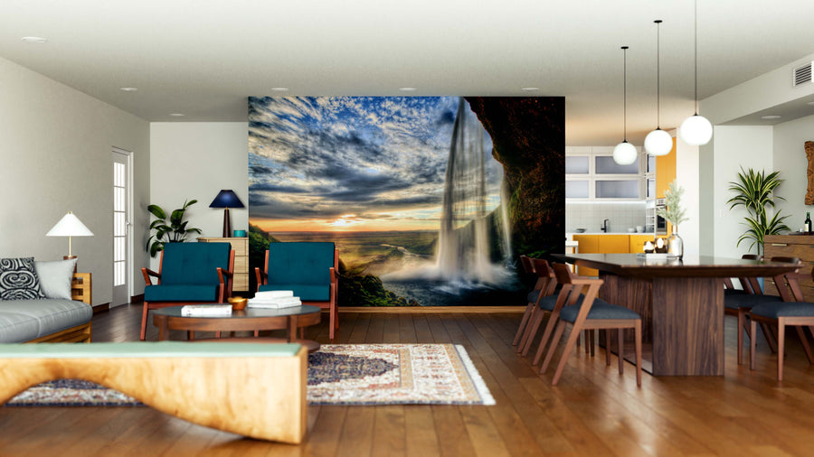 A modern open-concept living and dining room features wood floors, dark blue armchairs, a long dining table, and the Seljalandfoss Waterfall at Sunset Wall Mural as a dramatic focal point in this bright, stylish space.