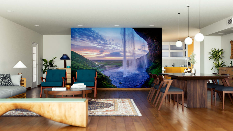 A modern open living-dining area features wood floors, blue and wood furniture, and a Seljalandfoss Waterfalls Wall Mural at sunset for natural beauty. Pendant lights hang over the dining table; kitchen visible in the background.