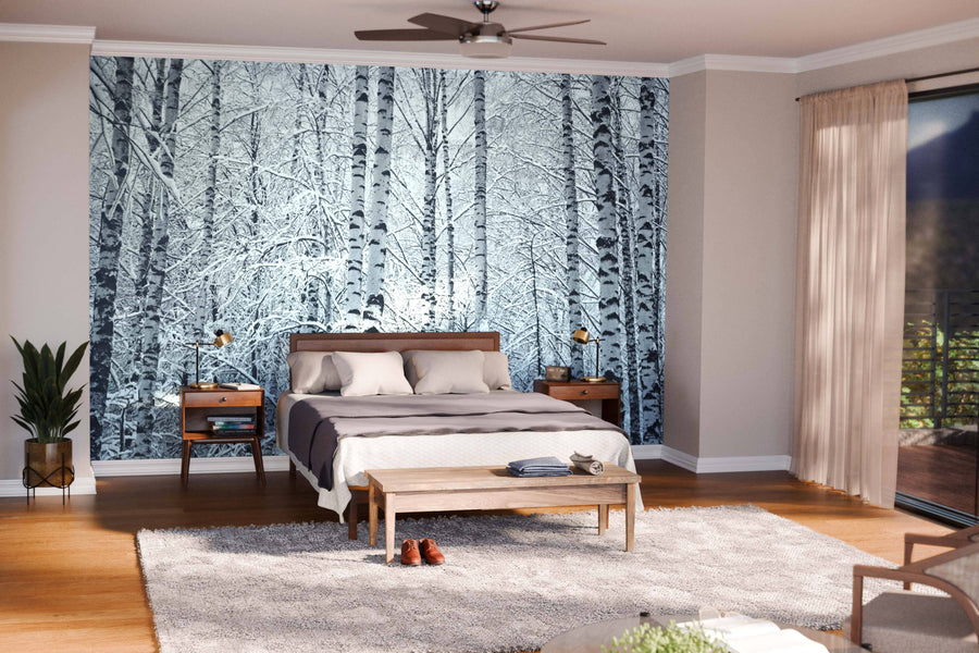 A modern open-plan living and dining area with wood floors, teal armchairs, a large table, and a decorative rug is accented by the Snow on the Branches Wall Mural, bringing a striking black-and-white birch tree design to the cozy interior.