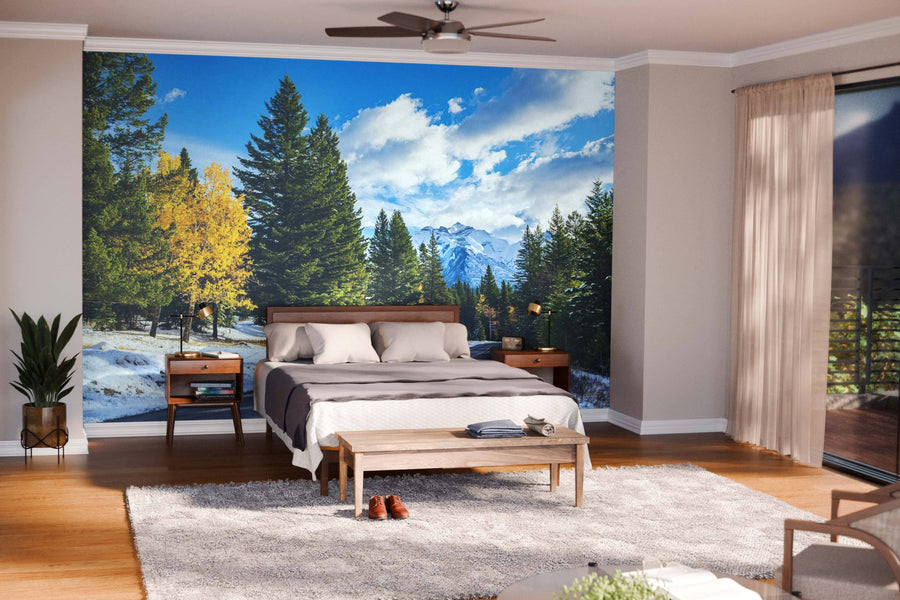 A modern living and dining room features wood floors, stylish furniture, and a Snowy Autumn Colorado Mountains Road Wall Mural. Warm lights, rugs, and pendant lamps add coziness, with the kitchen visible in the background.