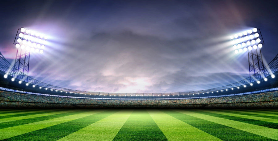 A bedroom decorated with the Stadium Glow Wall Mural—depicting a brightly lit soccer stadium at dusk—features a bed, computer desk, chair, soccer ball, and modern decor. Natural light through blinds enhances the sporty ambiance.