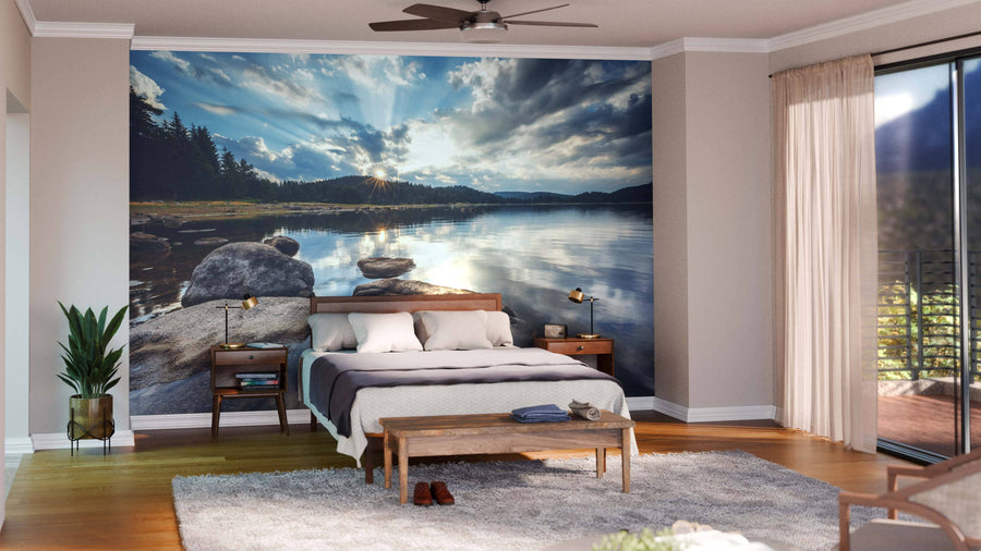 A modern bedroom features the Sun Rays Over A Mountain Lake Wall Mural behind a neatly made bed. The room has wood floors, a gray rug, plants, nightstands, and large windows with sheer curtains letting in natural light.