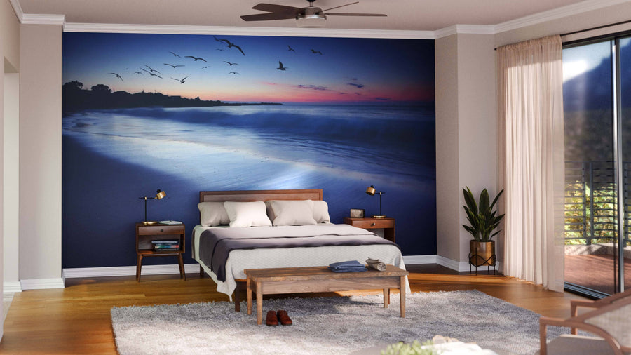 A modern living-dining room with wooden floors, teal chairs, a wooden bench, rugs, and the Sunset Beauty Wall Mural showing a beach sunset with birds. Pendant lights hang above a dark dining table, with a sideboard in the background.