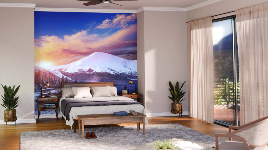 A modern living-dining area with wood floors, teal armchairs, and pendant lights features the Sunset Over Mountain Cottage Wall Mural, depicting a snow-capped mountain at sunset above a village and adding color to the neutral space.