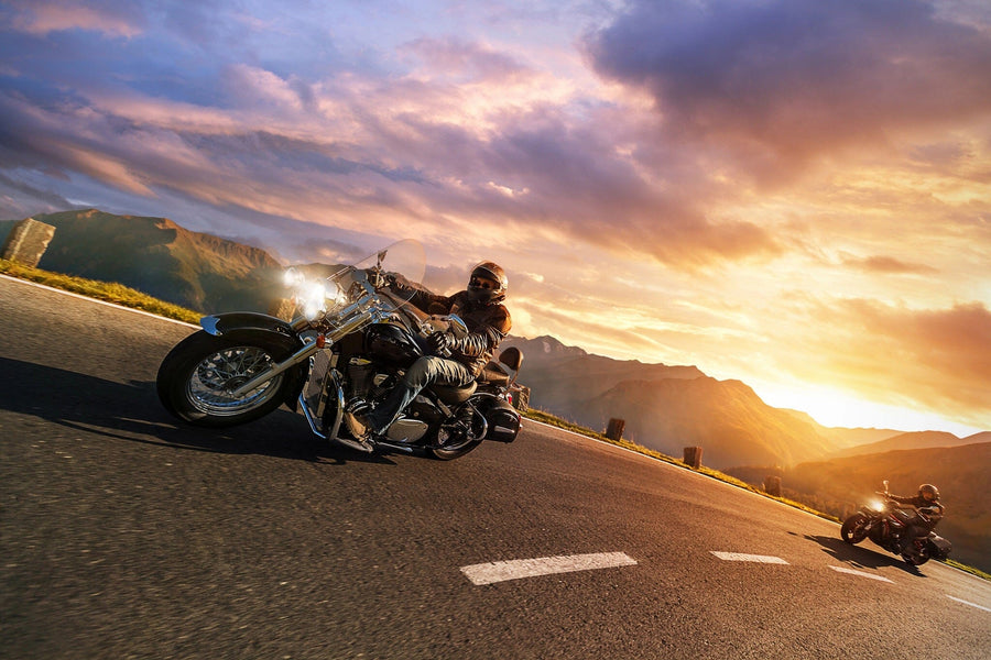 A modern garage with black cabinets, wall-mounted tools, and a retro motorcycle features the Sunset Ride Wall Mural on its back wall—depicting motorcyclists riding on a scenic mountain road at sunset.