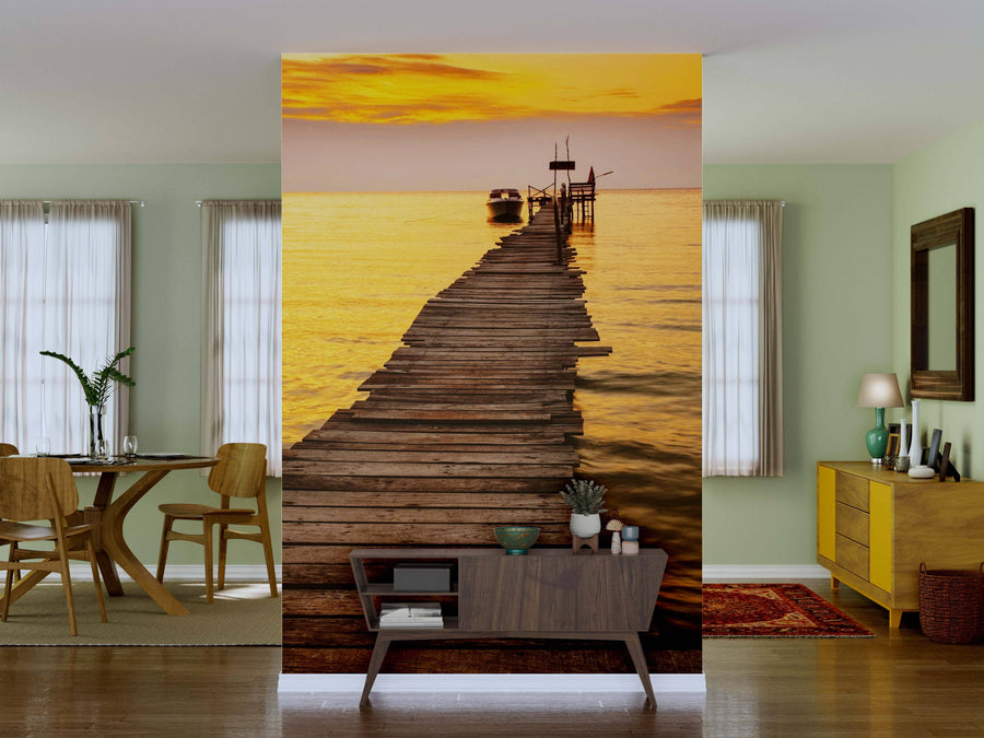 A modern living room with light green walls features the "Sunset on the Pier Wall Mural," showcasing a wooden pier at sunset over tranquil water. Mid-century furniture and plants add warmth and style to the inviting space.
