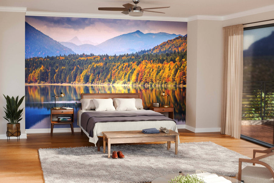 A cozy bedroom features the Sylvenstein Lake in Bavarian Alps Wall Mural of a lake, autumn forest, and mountains. The room includes a wooden bed with beige bedding, bedside tables, indoor plants, a bench, and large windows letting in natural light.